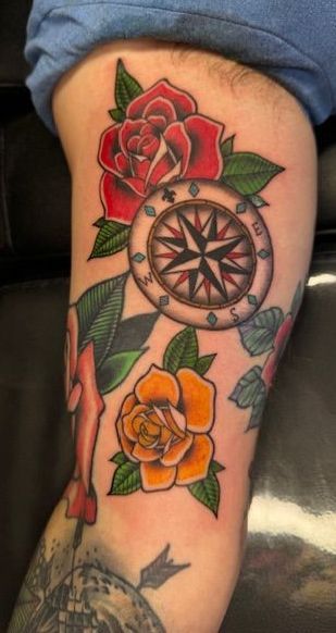 Forearm tattoo featuring red and orange roses, a compass, and leafy green accents