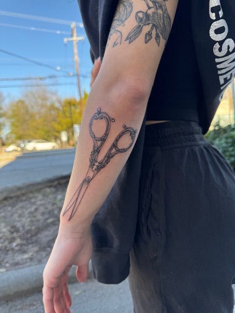 Person’s arm with black line-art tattoo, wearing a black sleeveless top, standing outdoors near a road.