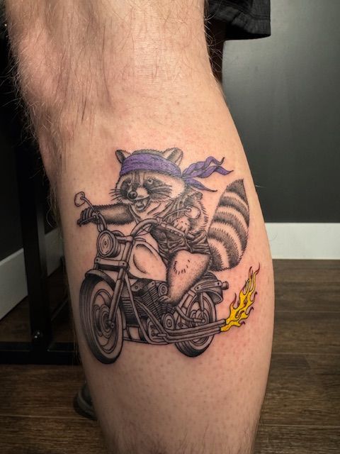 Black-and-white tattoo of a raccoon riding a motorcycle on a leg, with a small yellow flame accent