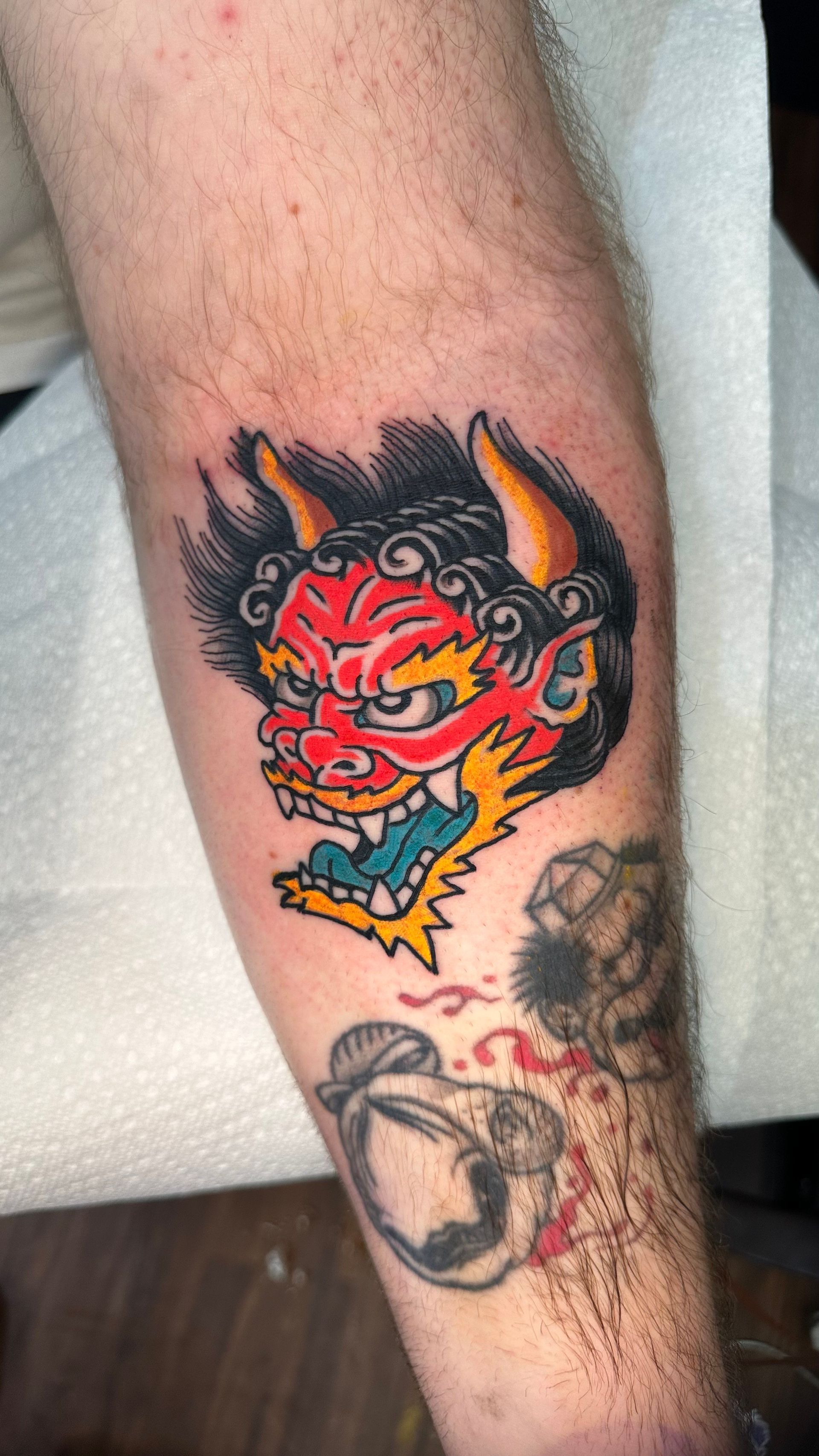Colorful red-and-yellow oni tattoo on a forearm, with a faded black sketch tattoo below.