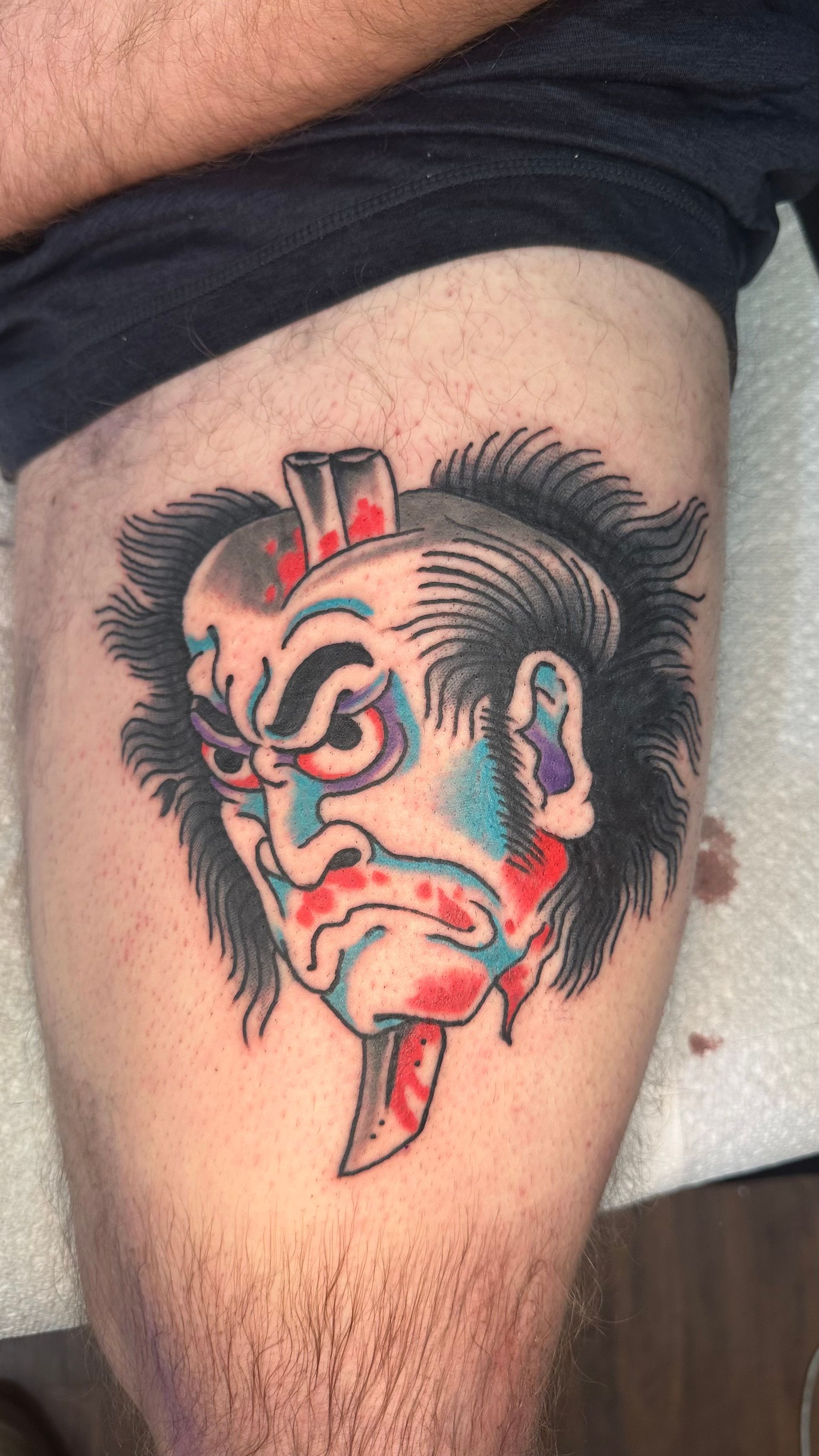 Colorful red, blue, and black oni mask tattoo on a hairy thigh