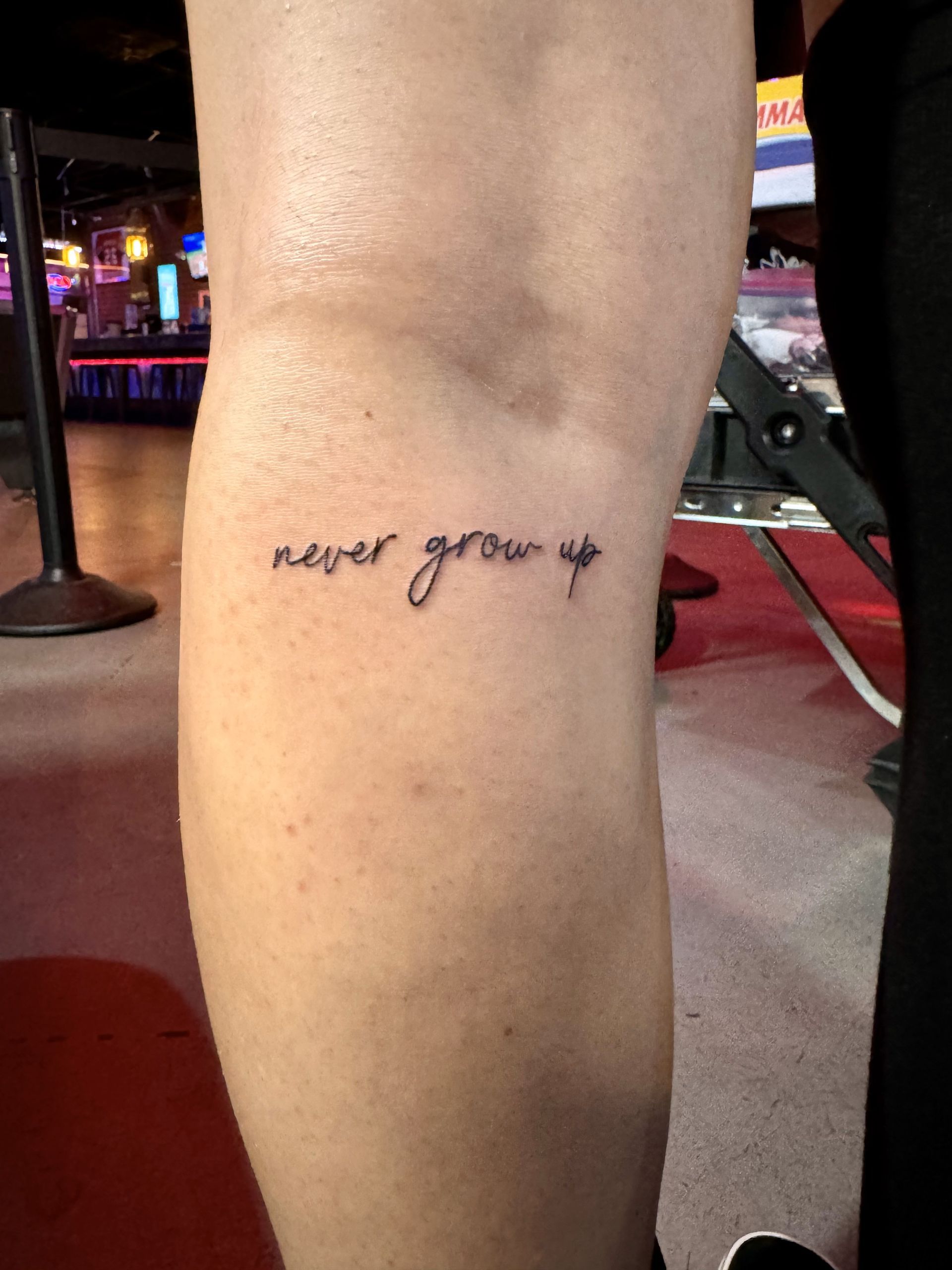 Close-up of a leg with a small “never grow up” tattoo on a red-carpeted walkway at night.