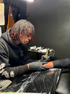Tattoo artist smiling while tattooing a client in a shop, wearing black gloves and holding a tattoo machine