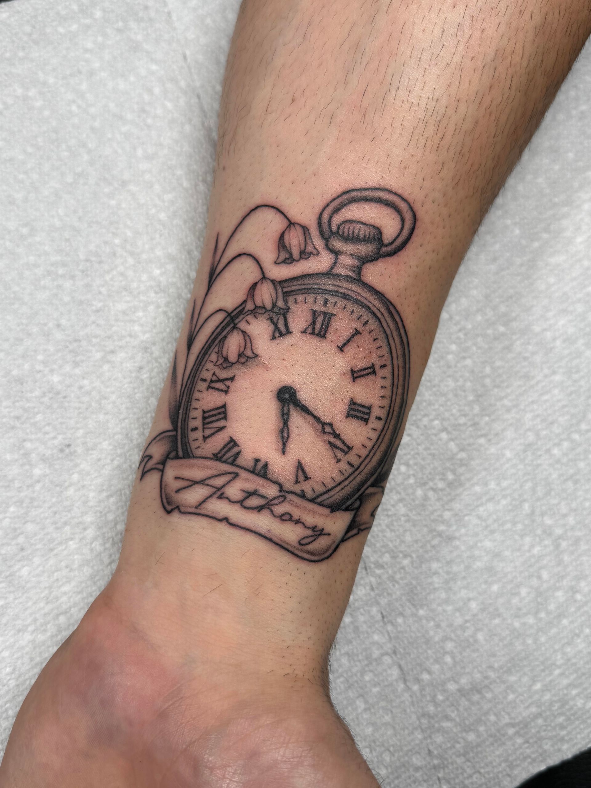 Black line-art tattoo of a pocket watch on a wrist, with a banner reading “Love.”