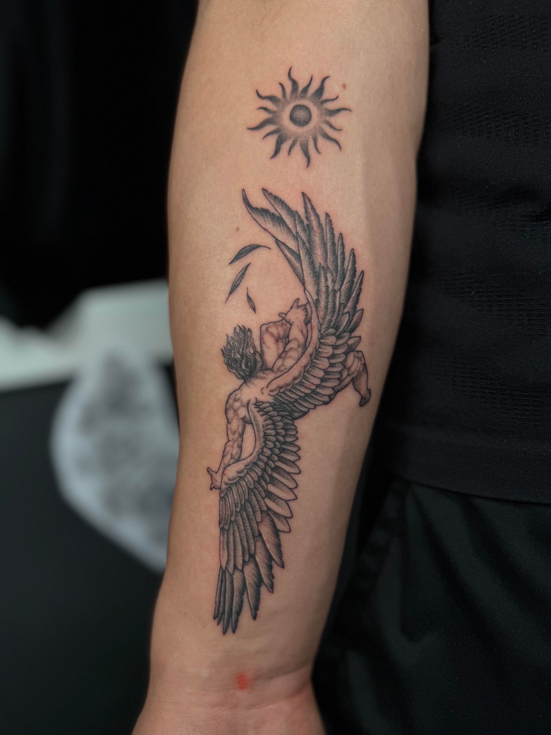 Arm tattoo of a winged angel beneath a small sun symbol on a person’s forearm