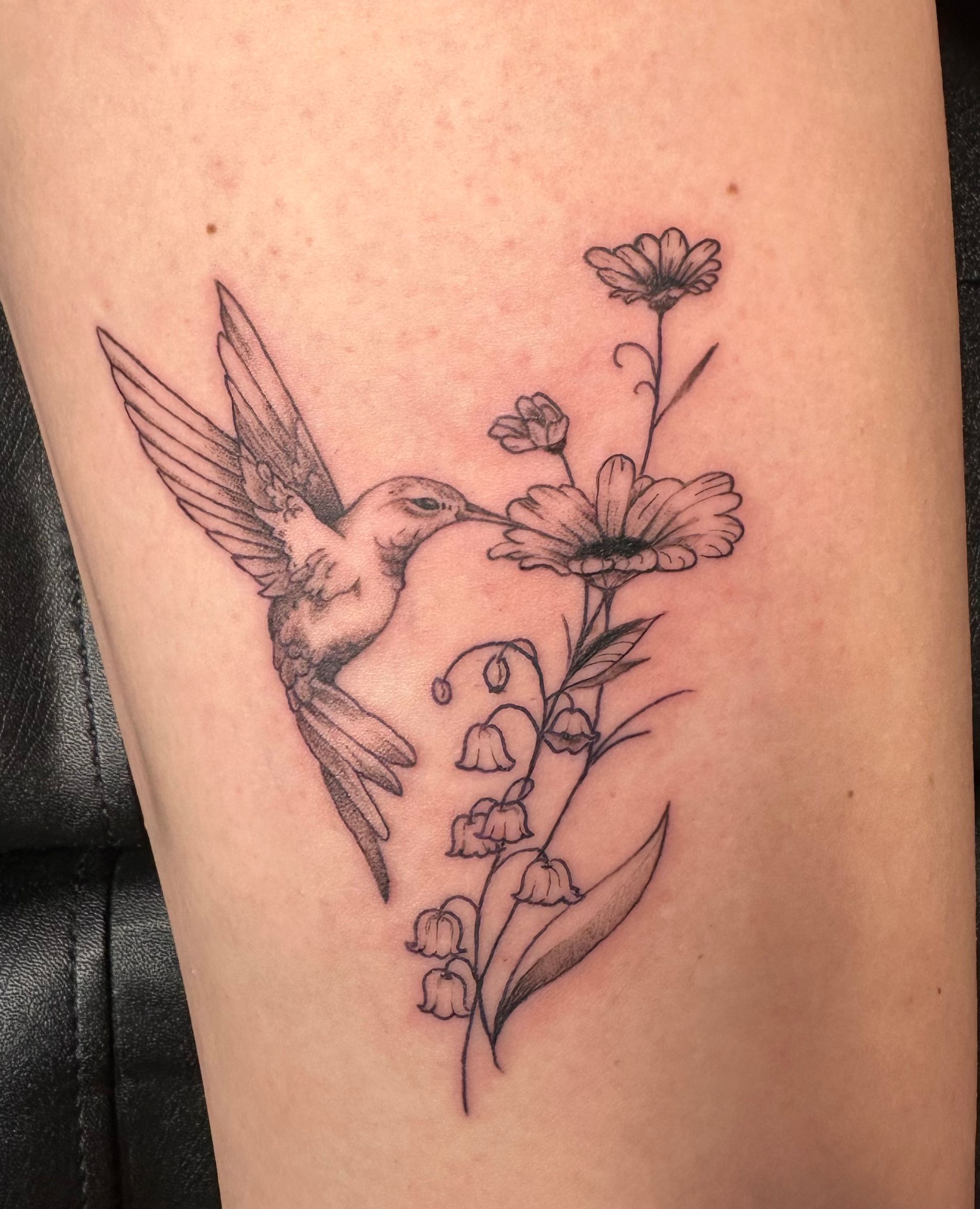 Black line tattoo of a hummingbird beside a flowering plant on skin