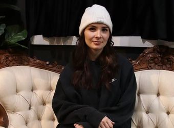 Woman in a white beanie and black sweater sitting on a cream tufted sofa, smiling softly.