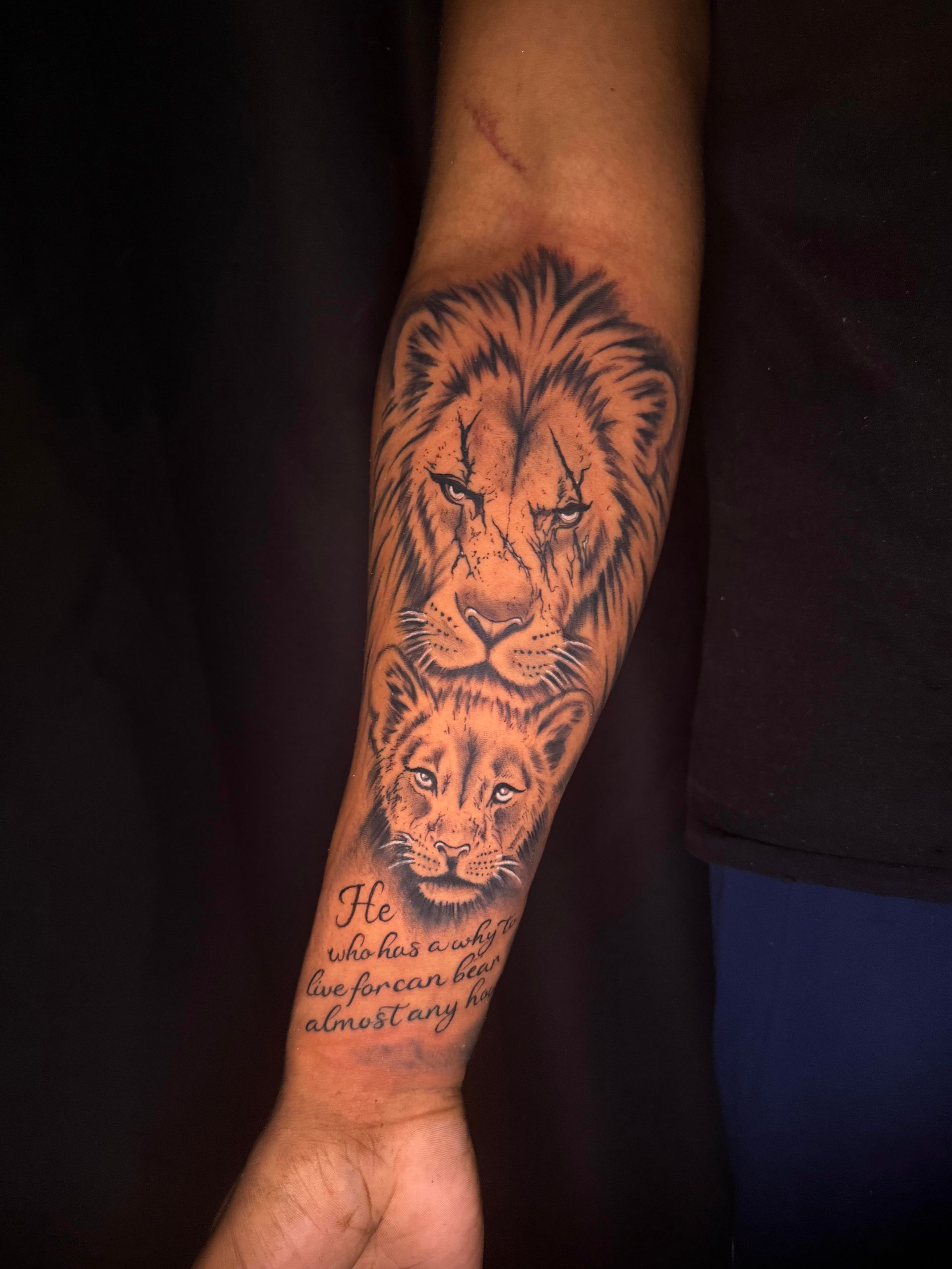 Forearm tattoo of a lion above a quote, in black ink on skin.