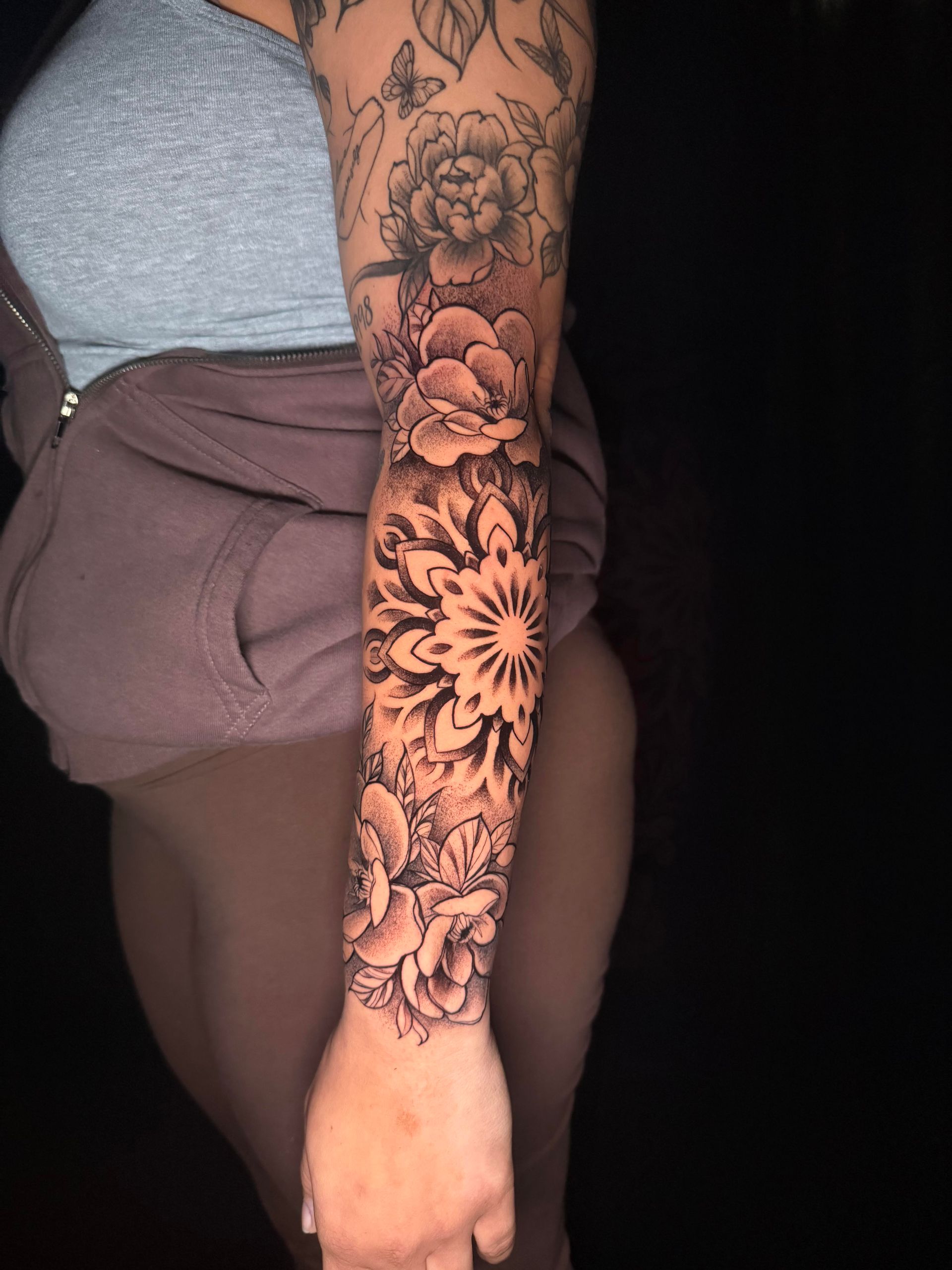 Arm with intricate black floral sleeve tattoo, hanging beside a gray shirt and mauve shorts.