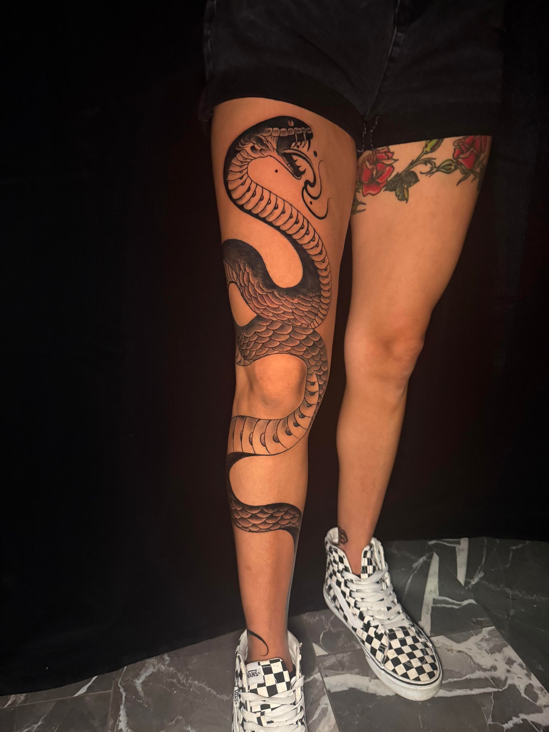 Person standing in black shorts, showcasing a large snake tattoo on one leg and a colorful thigh tattoo, wearing checkered sneakers