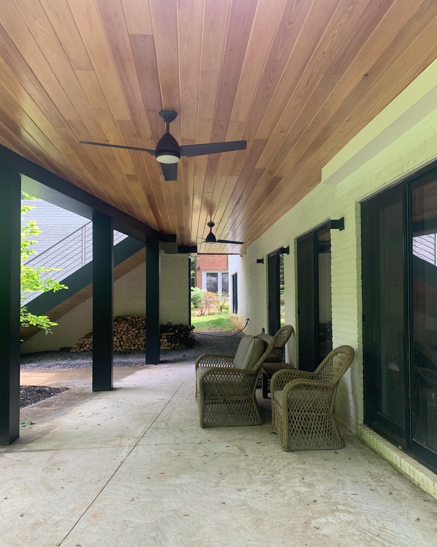 Covered porch with wicker chairs, ceiling fans, and dark support posts beside glass doors.