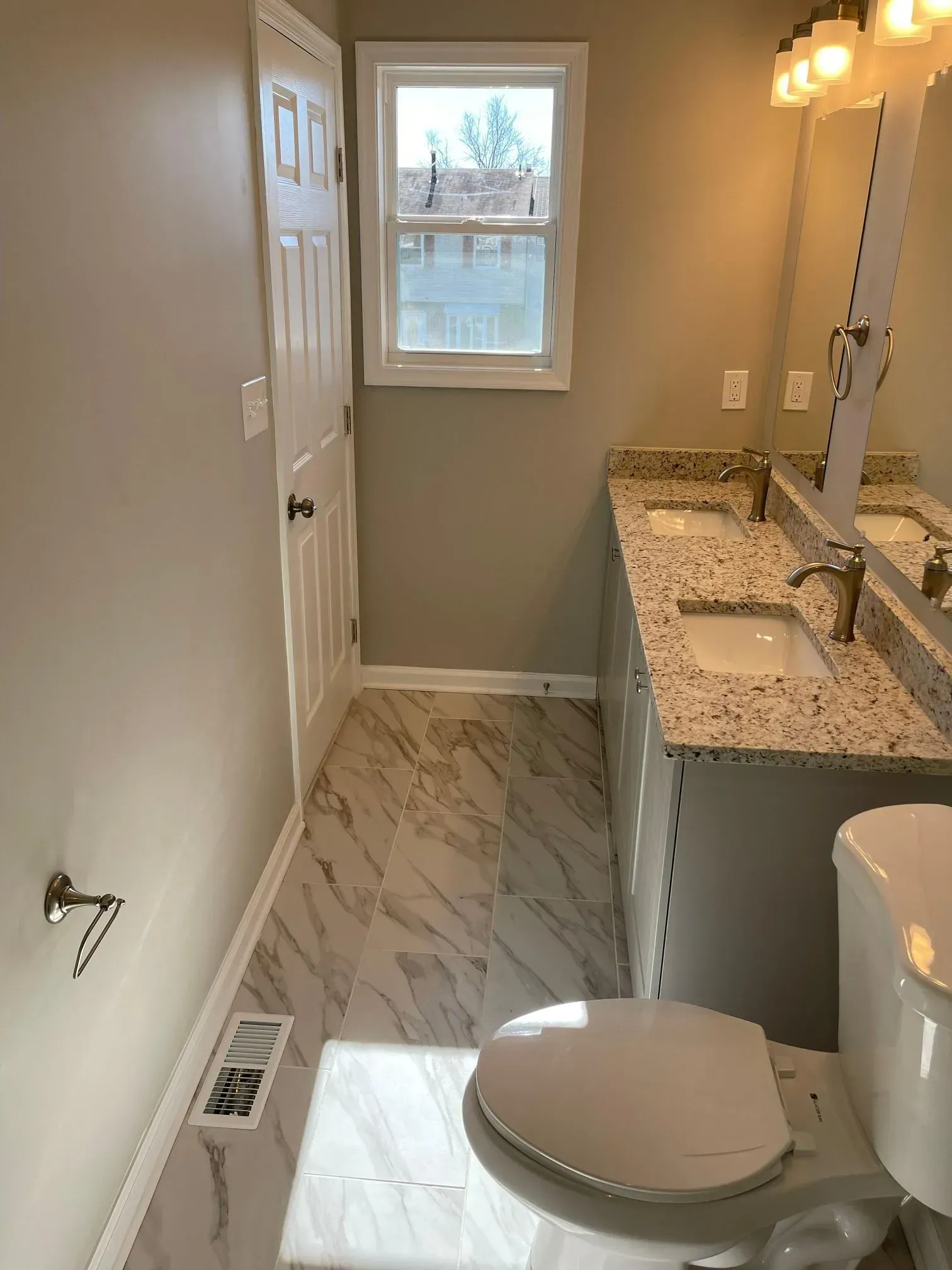 Narrow bathroom with white vanity, granite countertop, toilet, marble-look floor, and a window at the far end