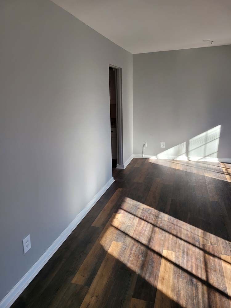 Empty room with gray walls, white trim, and dark wood-look flooring; sunlight streams through a window.