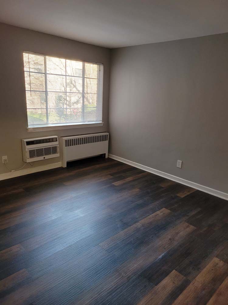 Empty room with dark wood-look flooring, light gray walls, window, and wall-mounted heating and cooling units.