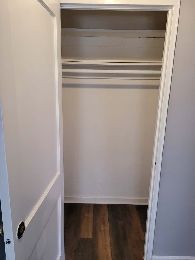 Empty white closet with a rod and shelf, hardwood-style floor, and a partially open white door.