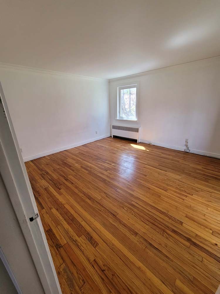 Empty room with hardwood floors, white walls, and a small window with radiator below.