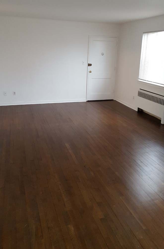 Empty room with wood flooring, white walls, and a door. There is a window with blinds and a radiator.