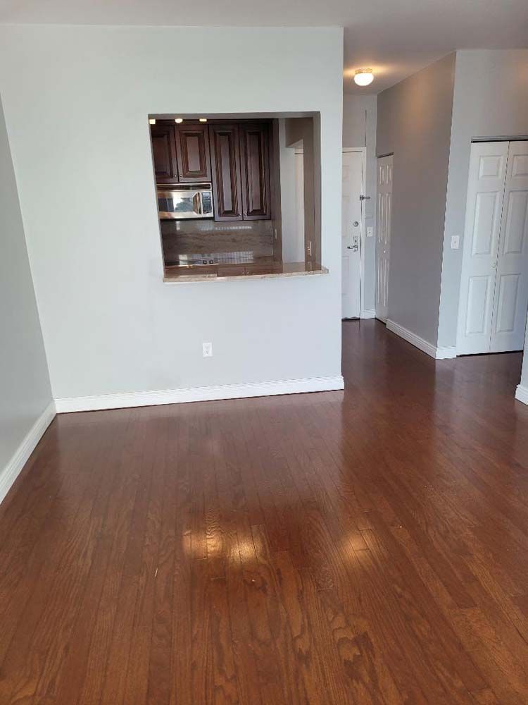 A room with wooden floors, gray walls, and a cutout looking into a kitchen with brown cabinets.