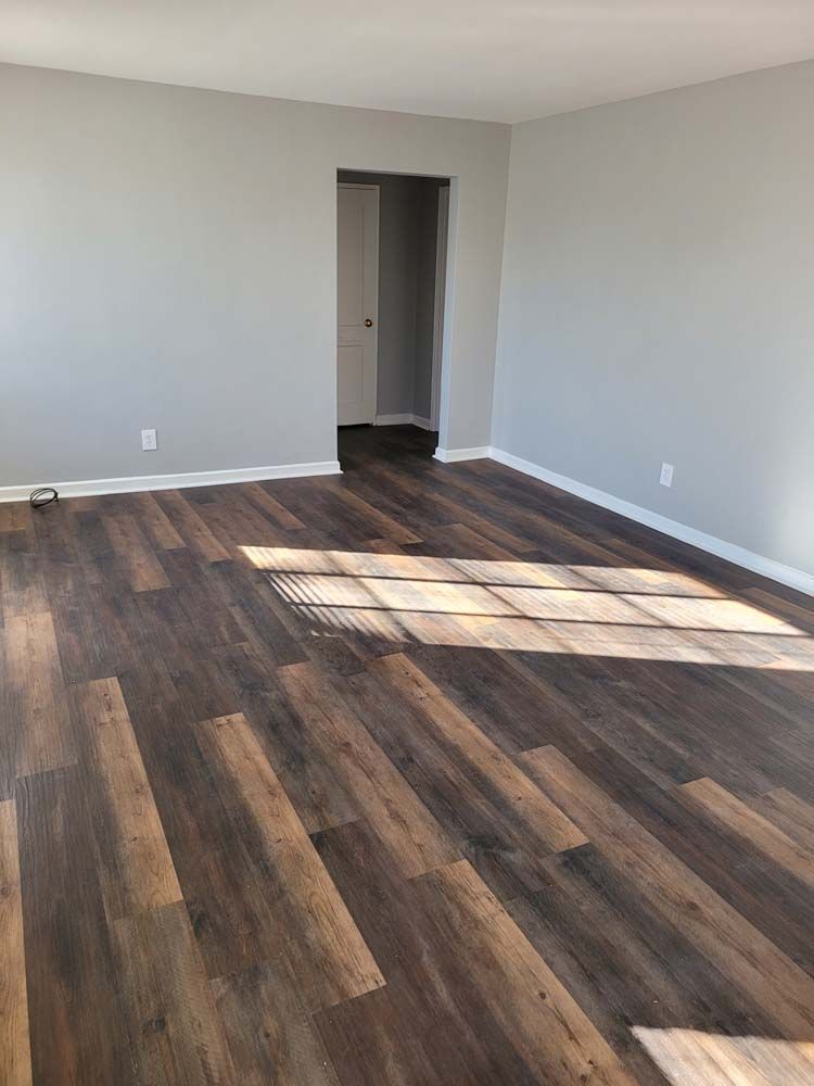 Empty room with gray walls, dark wood-look flooring, and a doorway. Sunlight streams across the floor.