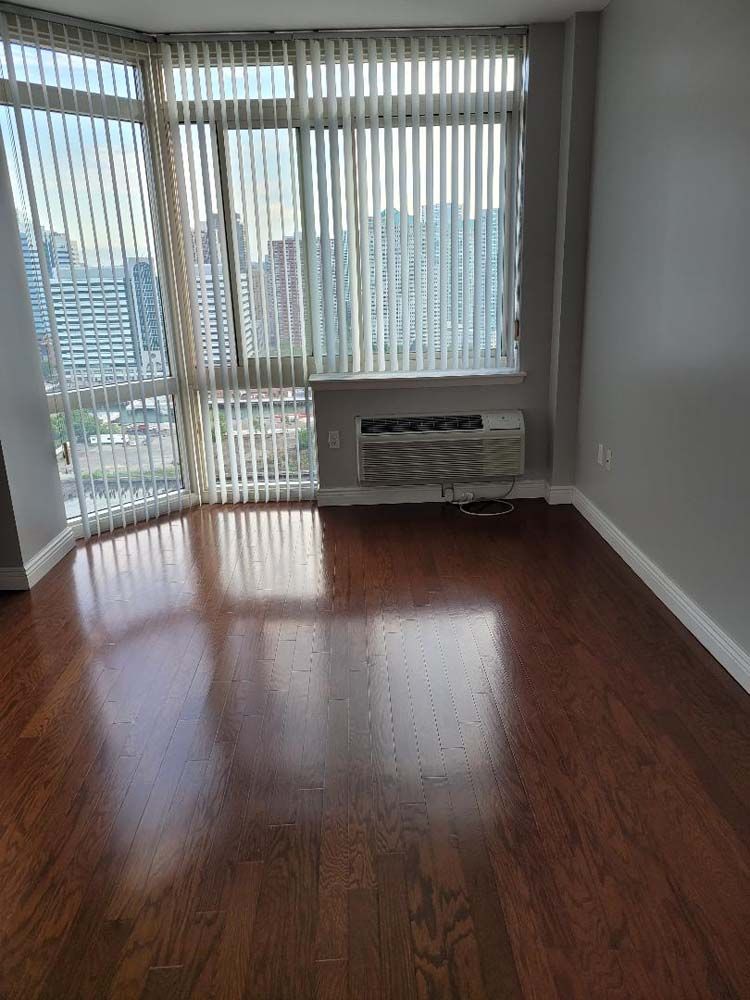 Empty room with hardwood floors, large windows with blinds, air conditioner, gray walls.