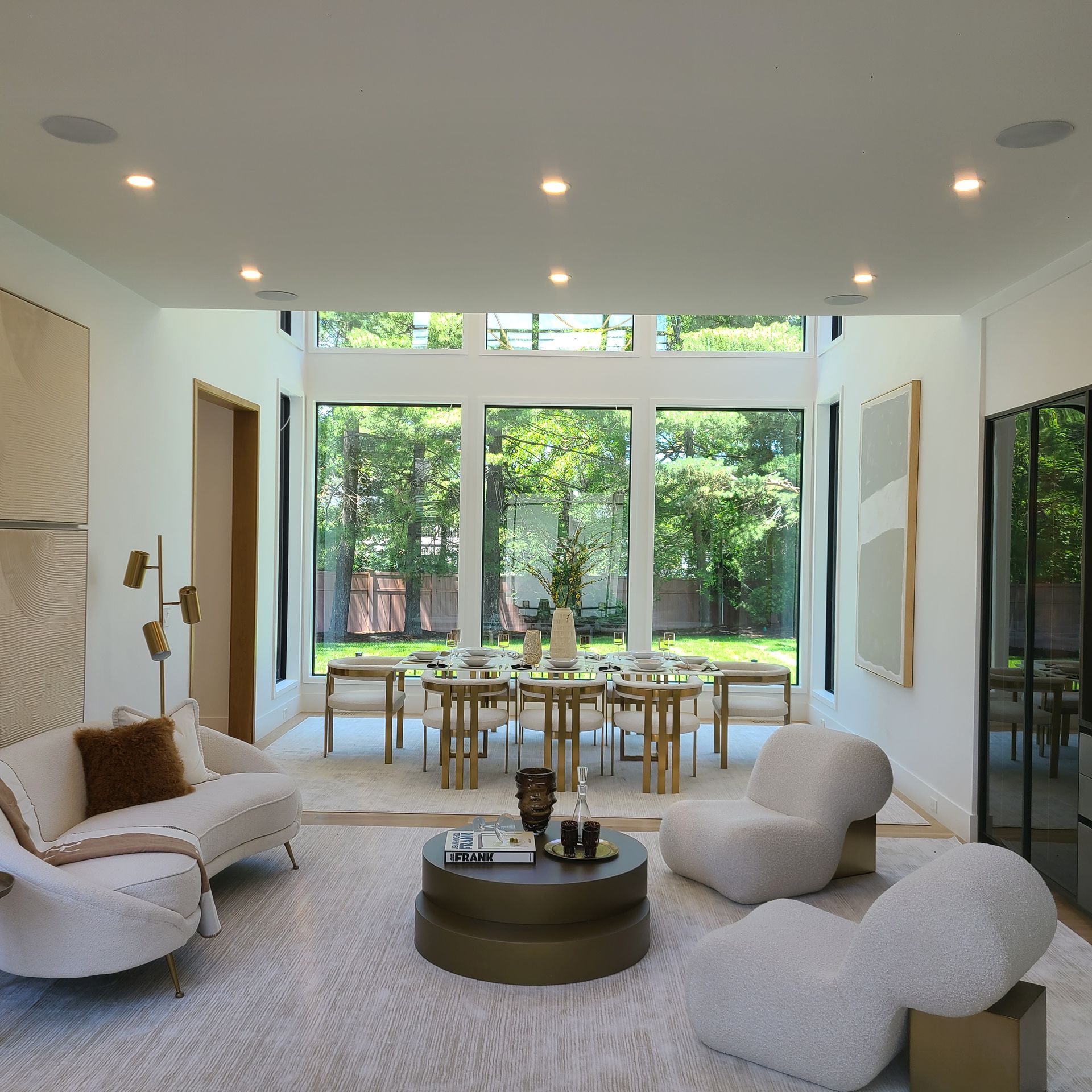 Modern living room with a dining table and chairs in front of large windows overlooking greenery.