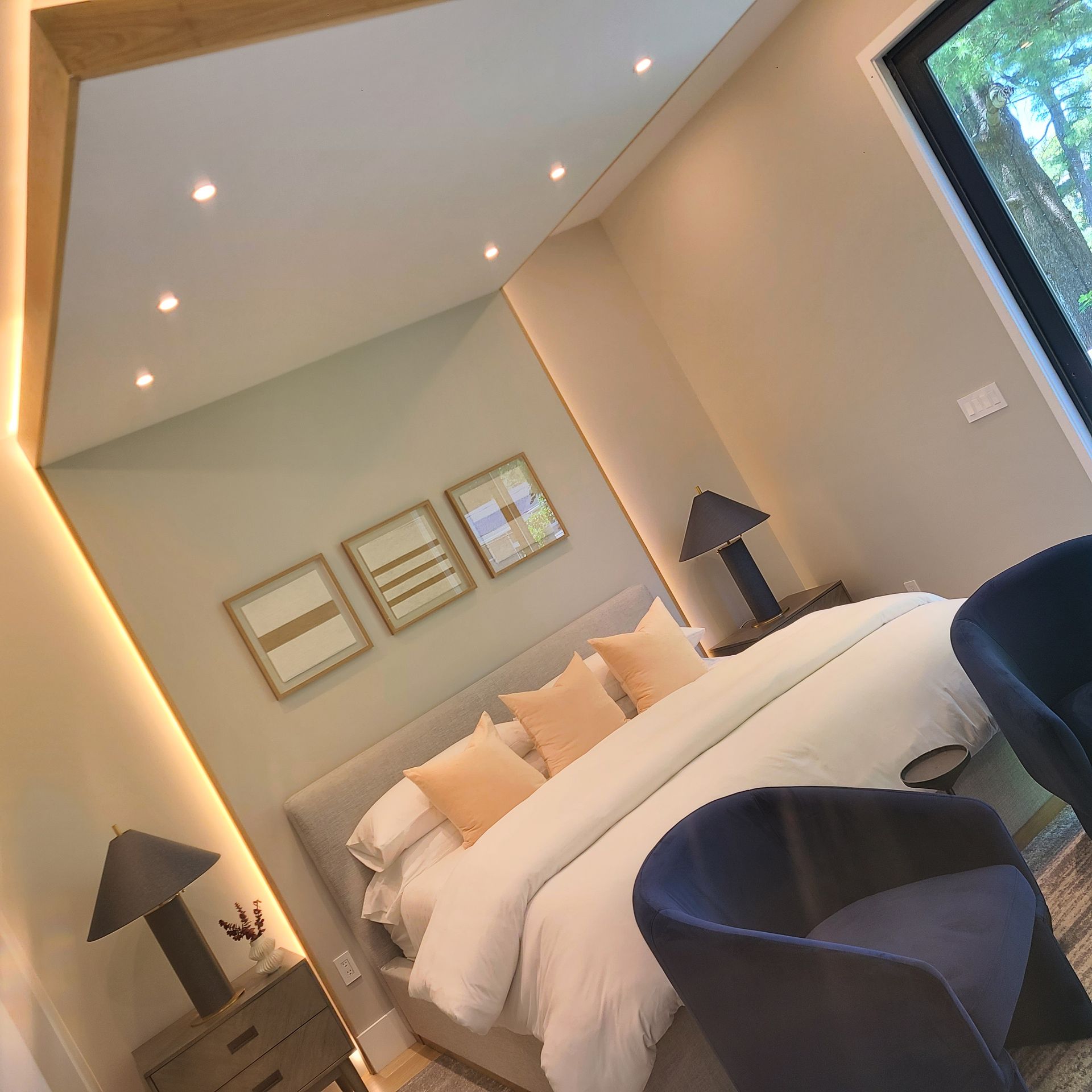 Bedroom with a bed, two chairs, and artwork. Neutral colors, accent lighting, and a window view.