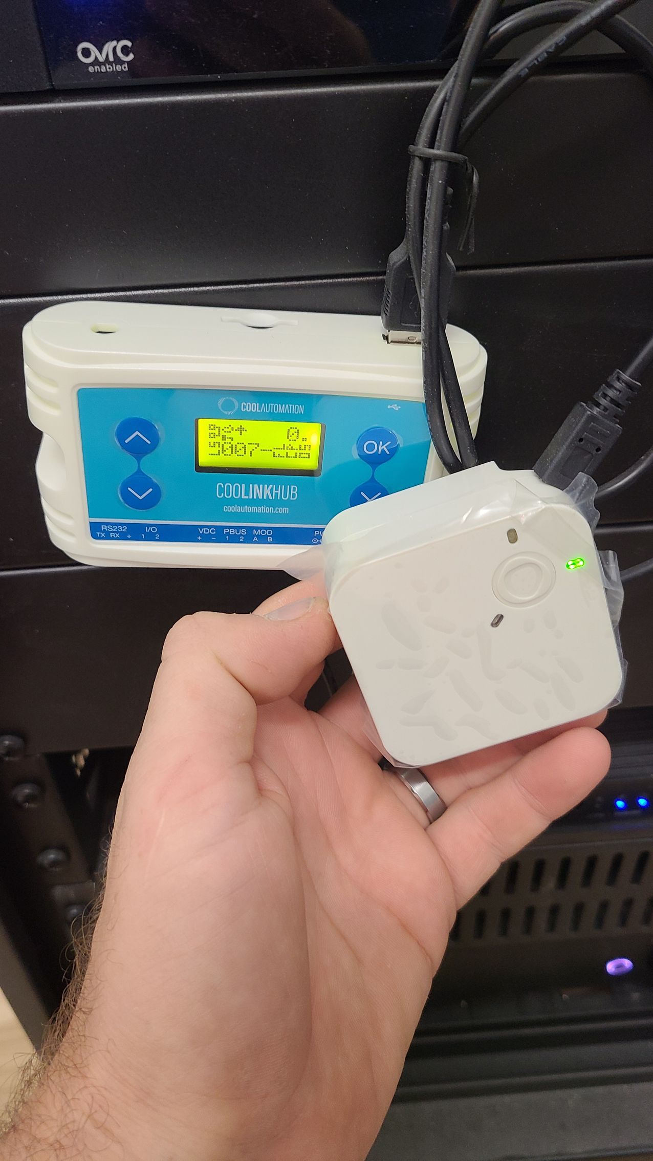 Hand holding a small white device with a green light, connected to a larger electronic device with a blue display screen and buttons.