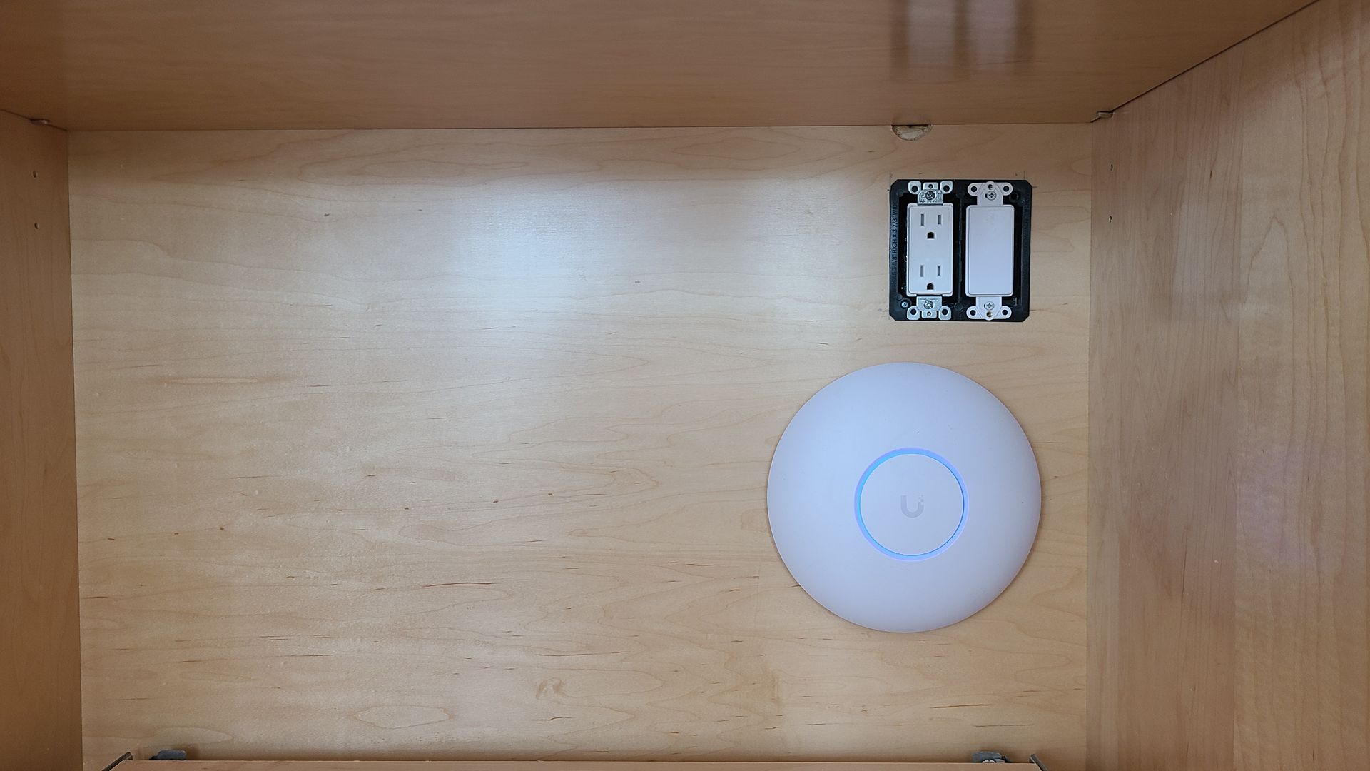Inside a wooden cabinet, a white Wi-Fi access point is mounted below a blank electrical outlet.