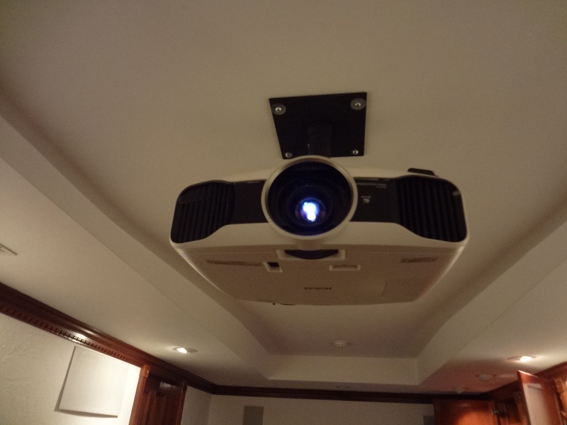 A white and black projector mounted on a white ceiling with a blue light shining.