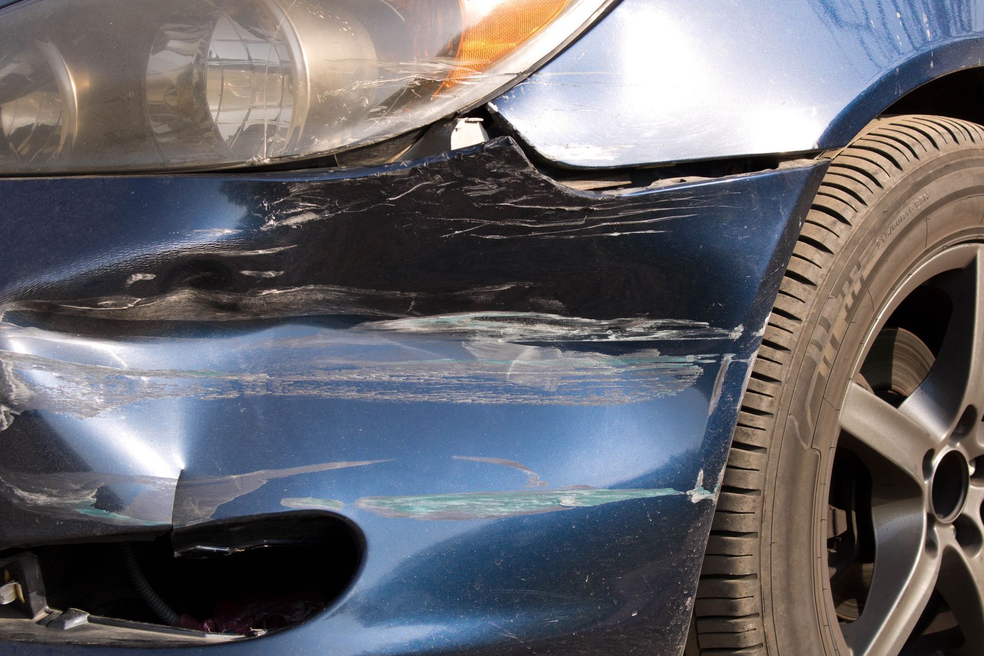Close-up of a blue car’s front bumper and fender with visible damage and scratches.