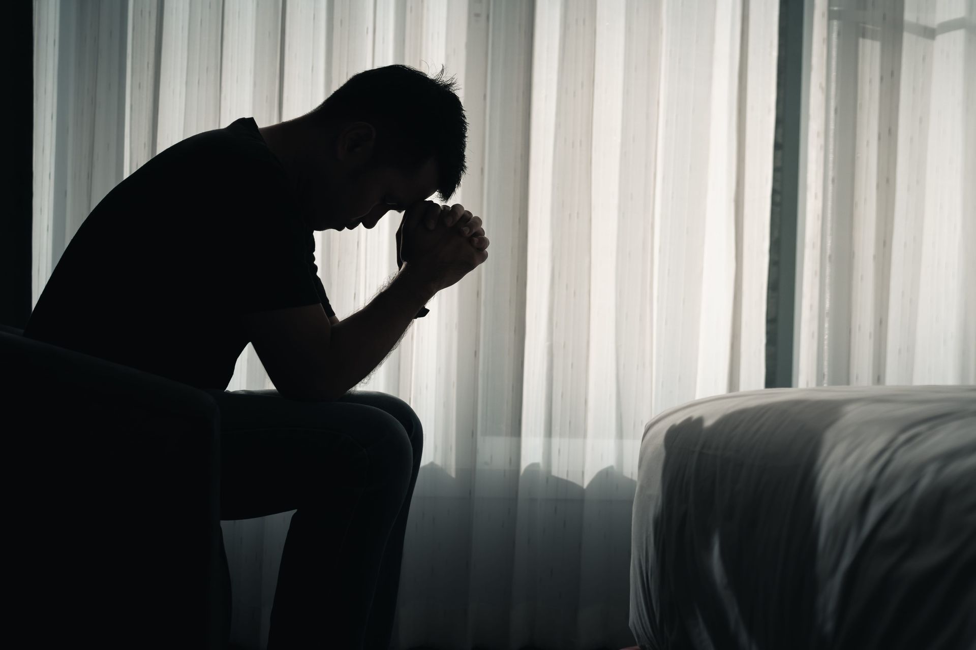 Silhouetted person sitting on a bed with head bowed in a dim room by a bright window