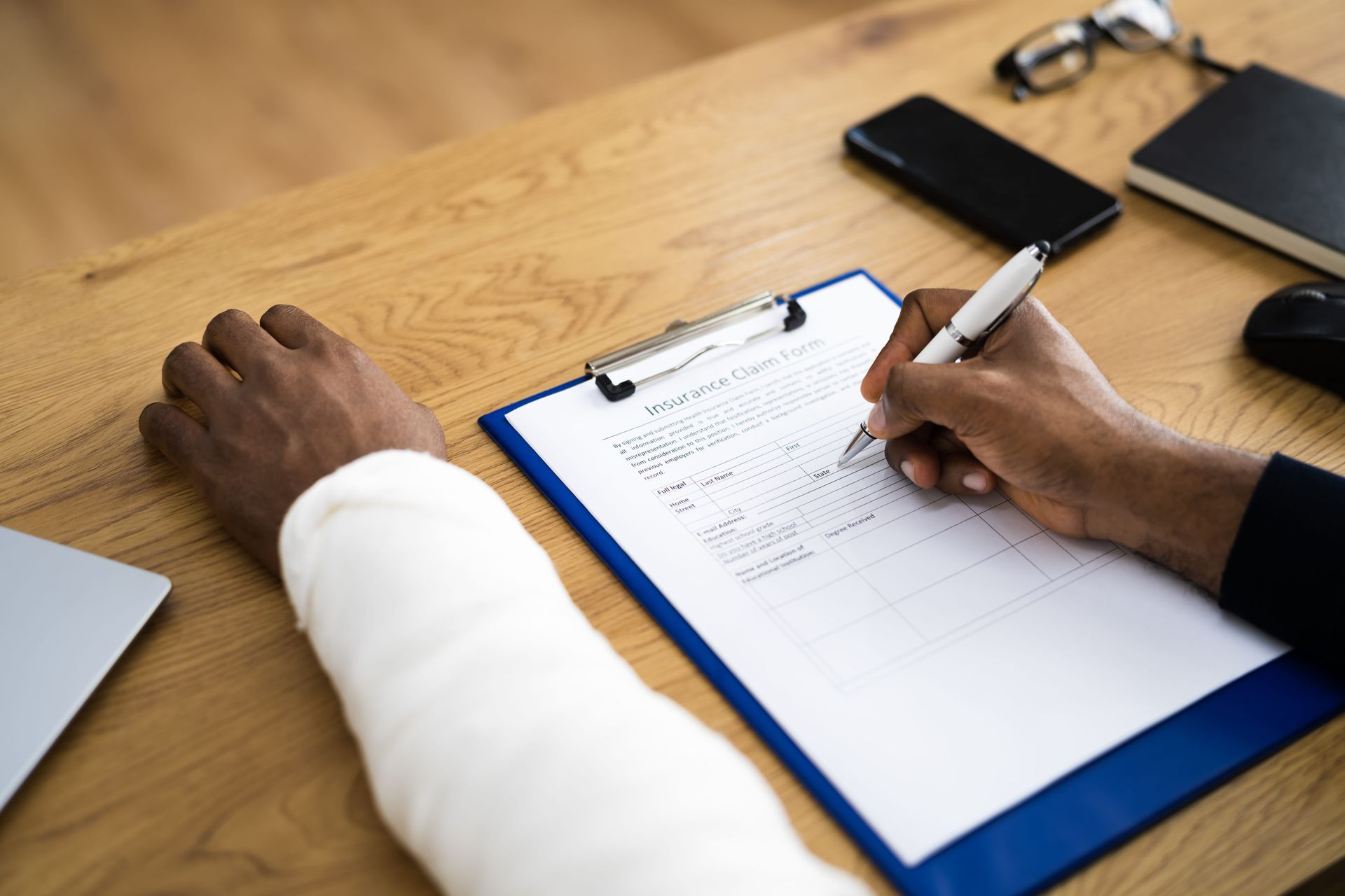 Person writing on a clipboard at a wooden desk with a phone and eyeglasses nearby.