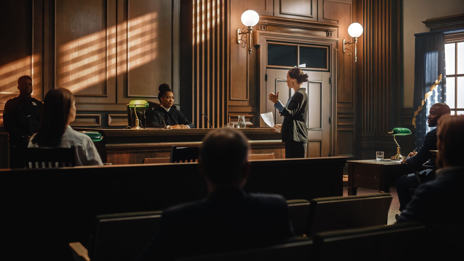 Courtroom with people seated and a speaker at the bench, lit by warm sunlight through tall windows.