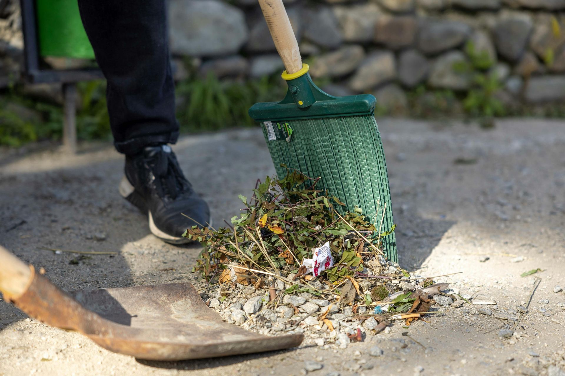 Two people using a green scoop and broom to collect leaves and debris from a paved path