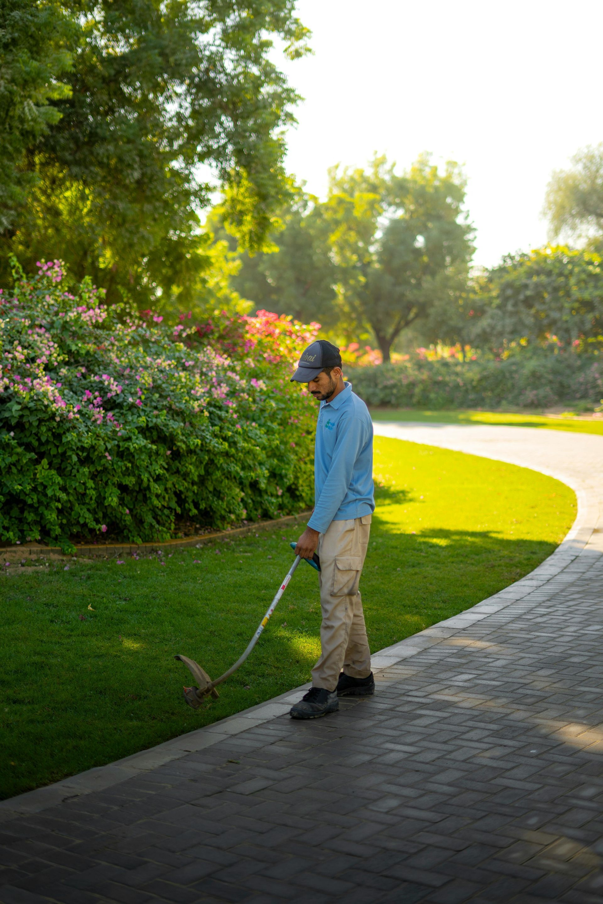 Person using a golf club on a paved path beside a sunlit garden with flowers and trees
