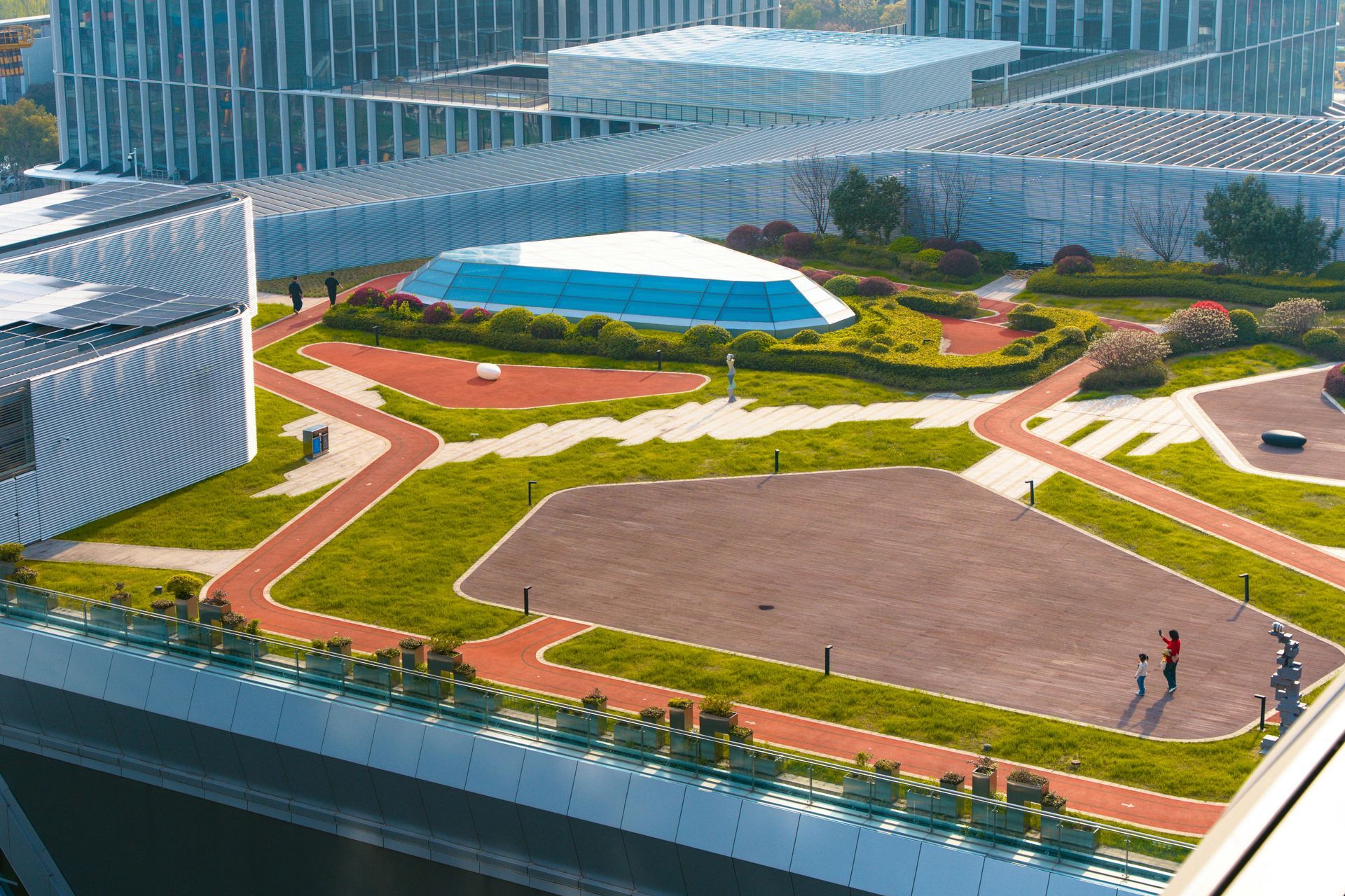 Modern rooftop park with winding paths, green lawns, and a blue-and-white pavilion among glass buildings