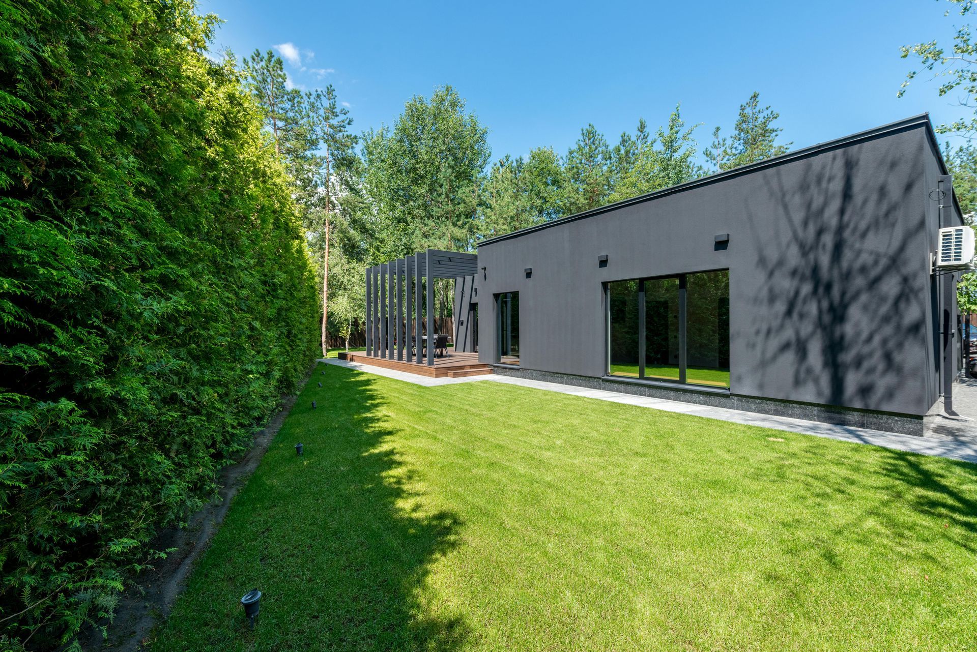 Modern gray house with large windows beside a green lawn and tall hedges under a bright blue sky