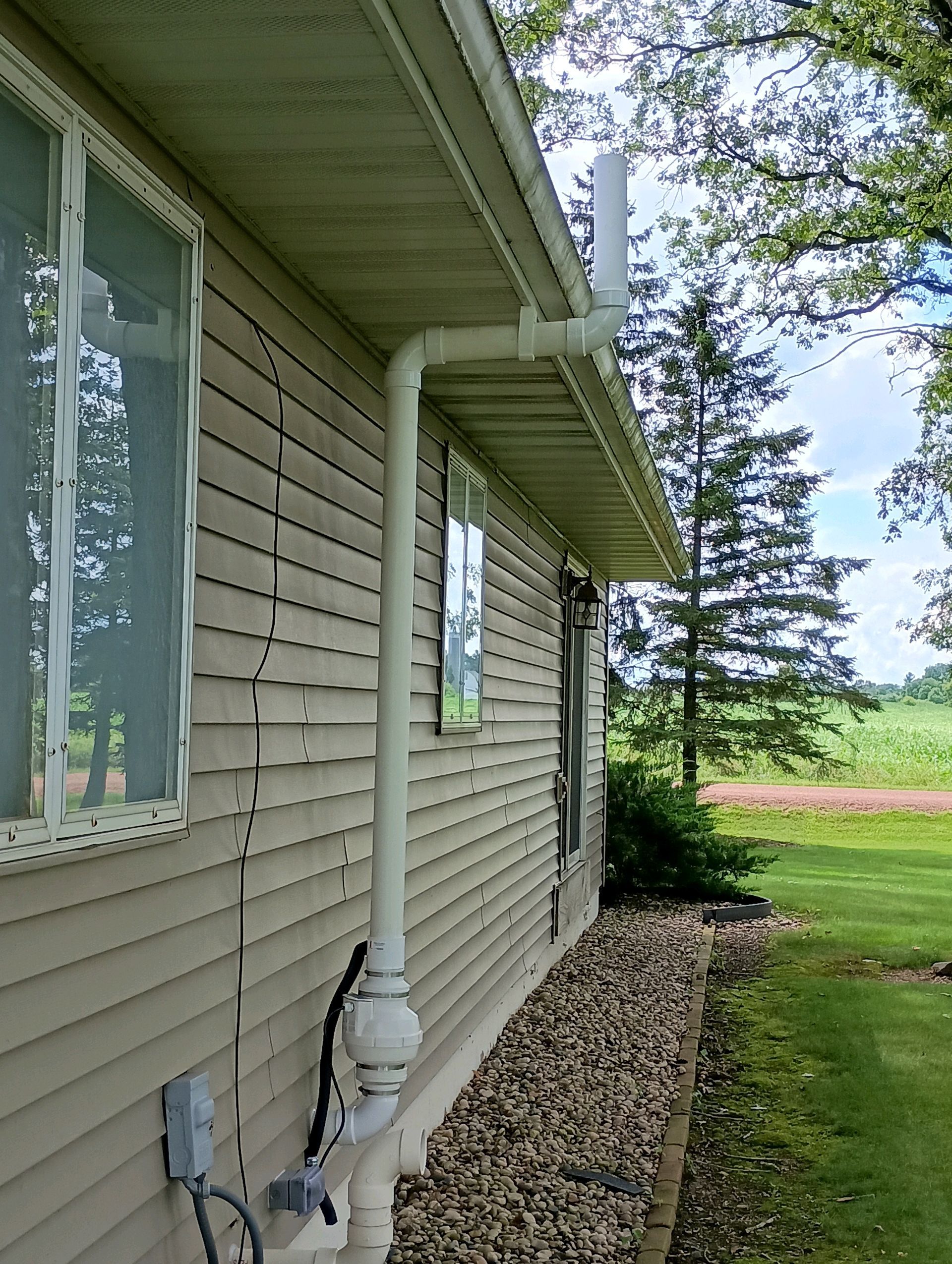 White pipe vent system on the side of a beige house with windows, rock border, and green field.