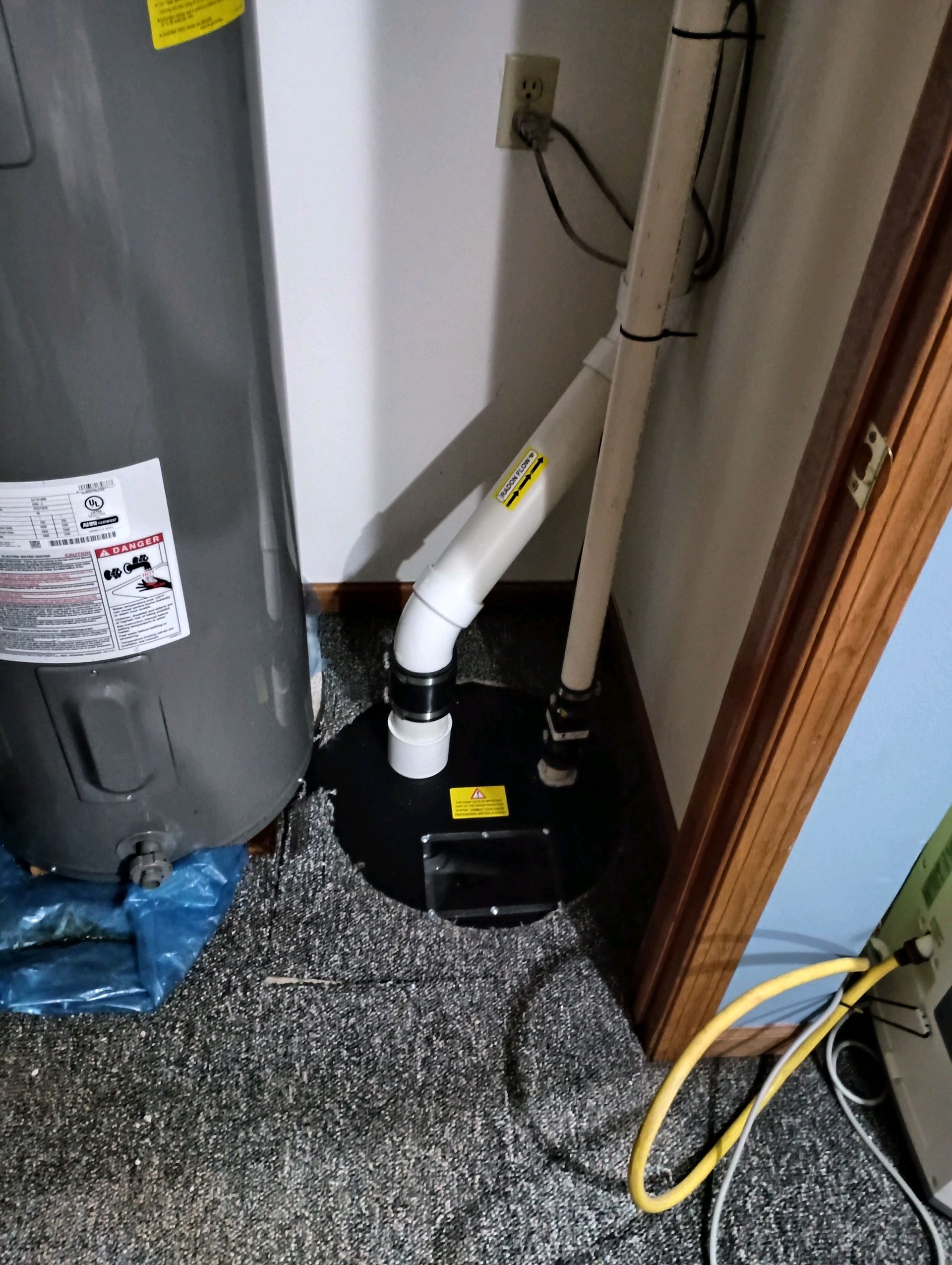 Sump pump installation in a corner near a water heater. White pipes, a black basin, and a gray carpet.