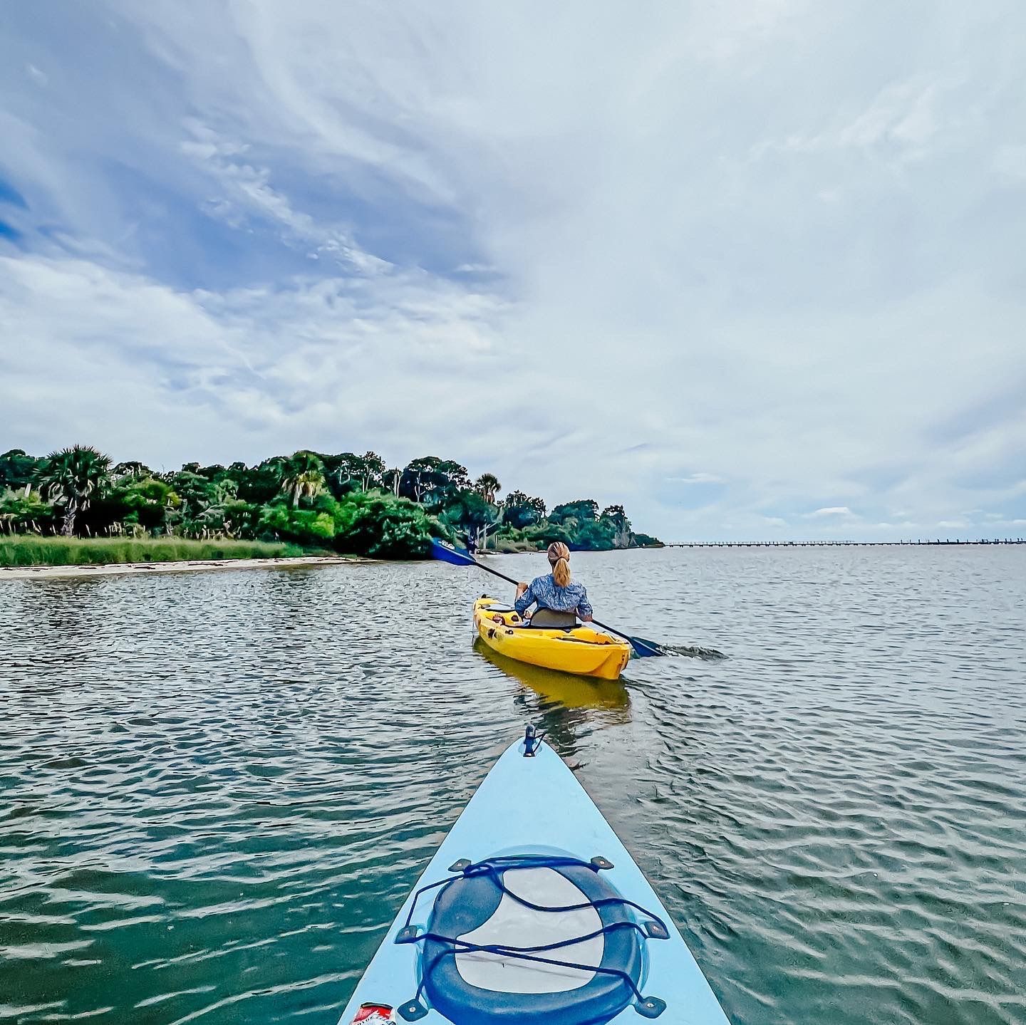 Kayaker in a yellow kayak on calm water, seen from a blue kayak near a tropical shoreline under cloudy sky