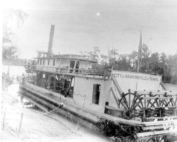 Black-and-white photo of a steamboat with paddle wheel docked on a riverbank.