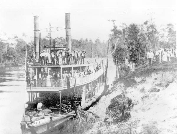 Vintage steamboat moving through a narrow canal beside trees and banks, with passengers on deck