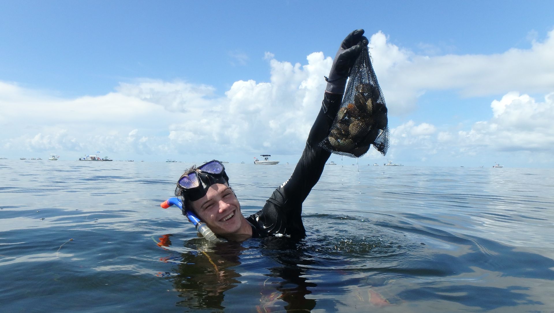 A snorkeler smiles while holding up a bag of scallops in calm open water