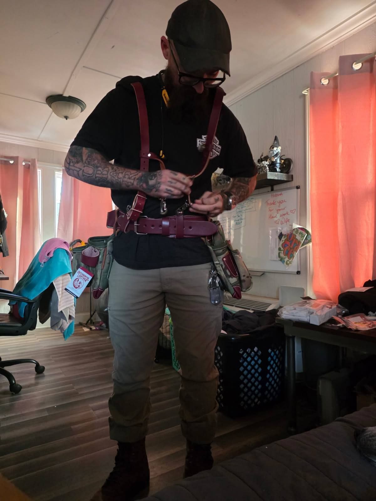 A person in a black t-shirt and tan pants adjusts a maroon leather utility belt and harness in a room with pink curtains.