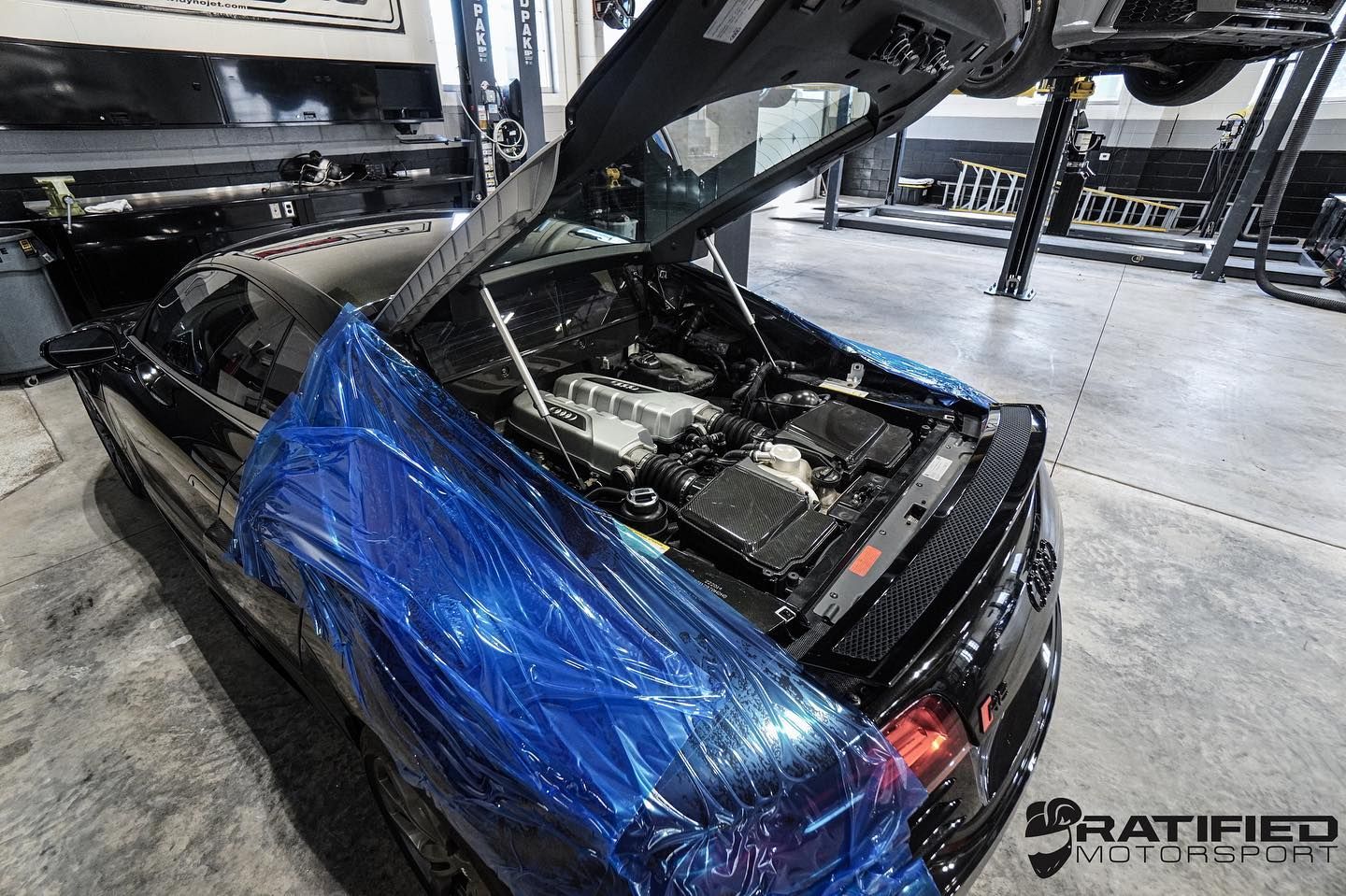 Black sports car with trunk open, blue protective film, engine visible, in a garage.