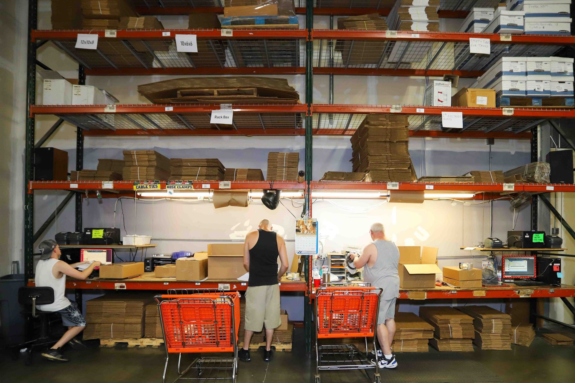 People packing orders at a warehouse workbench with shelves of boxes and bins in the background