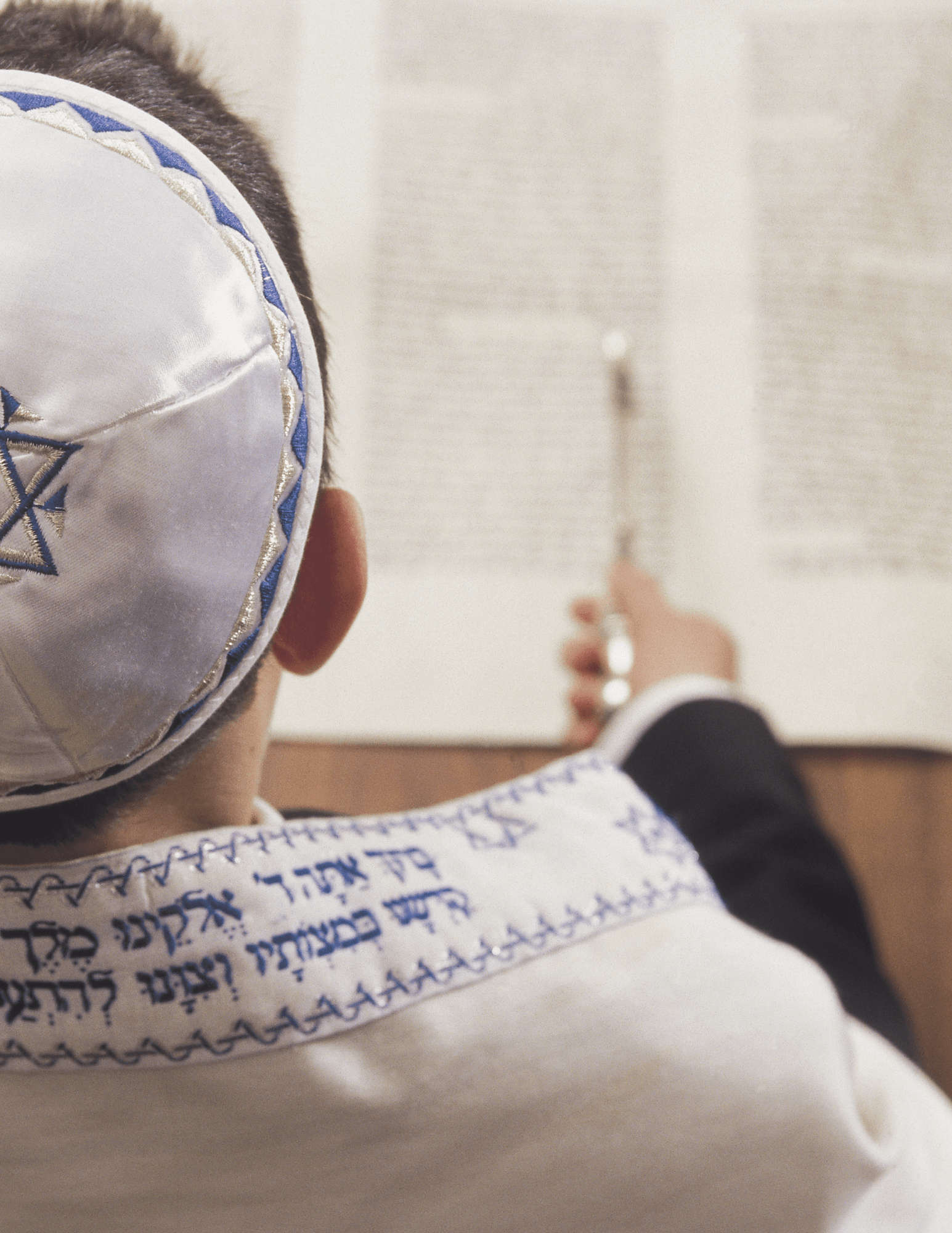 Photo of a b' mitzvah in kippah and tallit, their hand holding a yad to the Torah