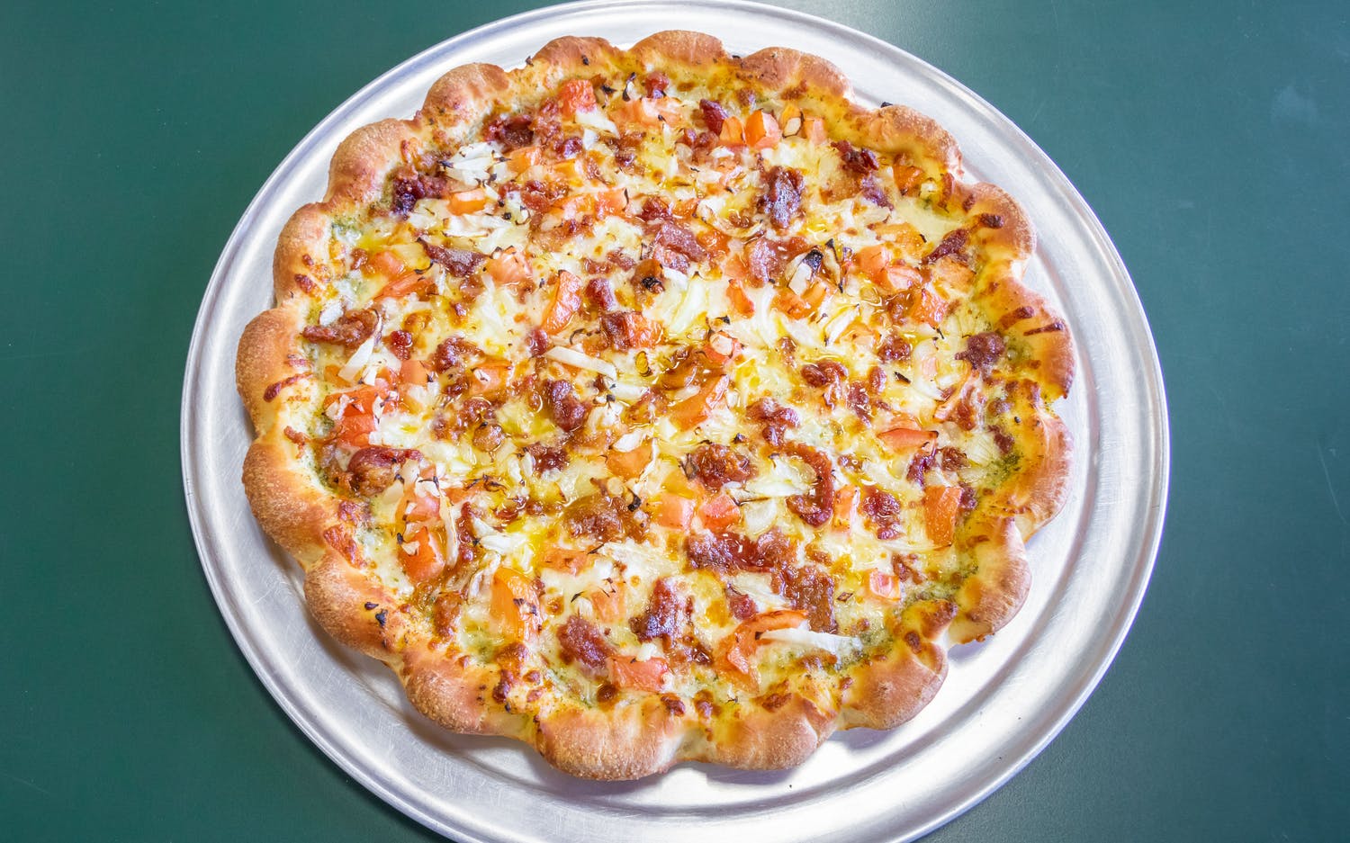 A pizza with a scalloped crust, topped with cheese, tomato, and bacon, resting on a round metal tray against a green table.