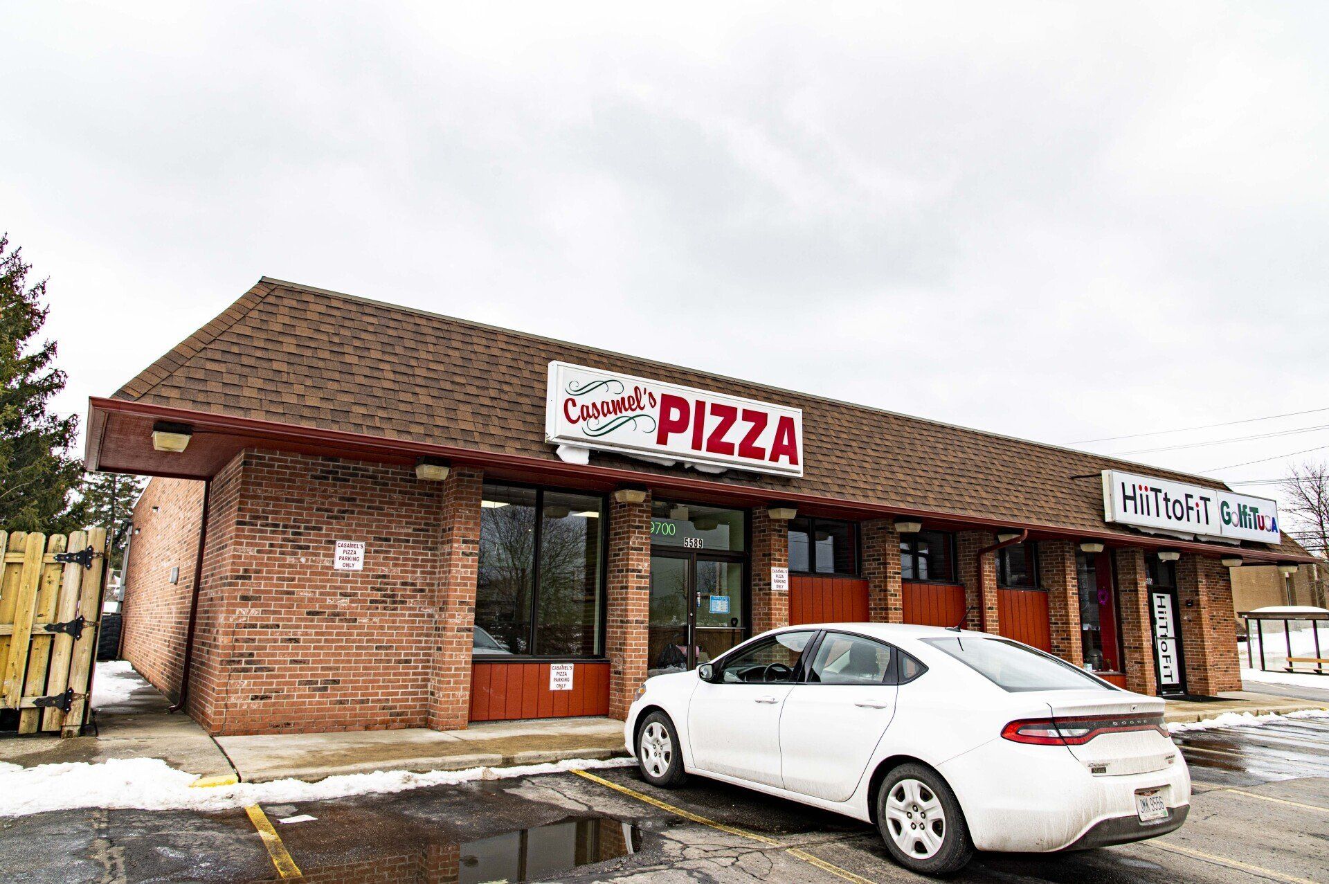 A brick pizza restaurant building with a white sedan parked in front on a cloudy day.