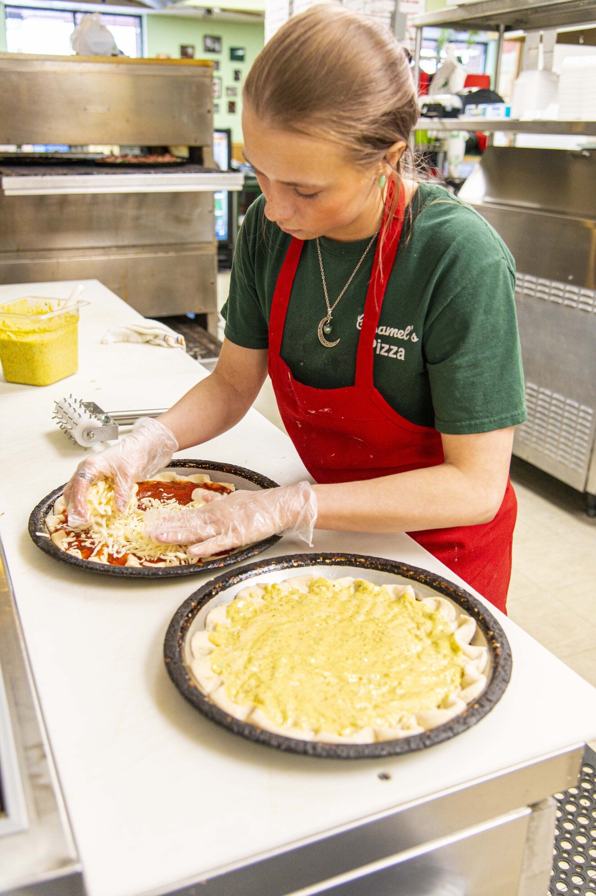 A food worker wearing a red apron and plastic gloves spreads cheese on a pizza inside a professional kitchen.